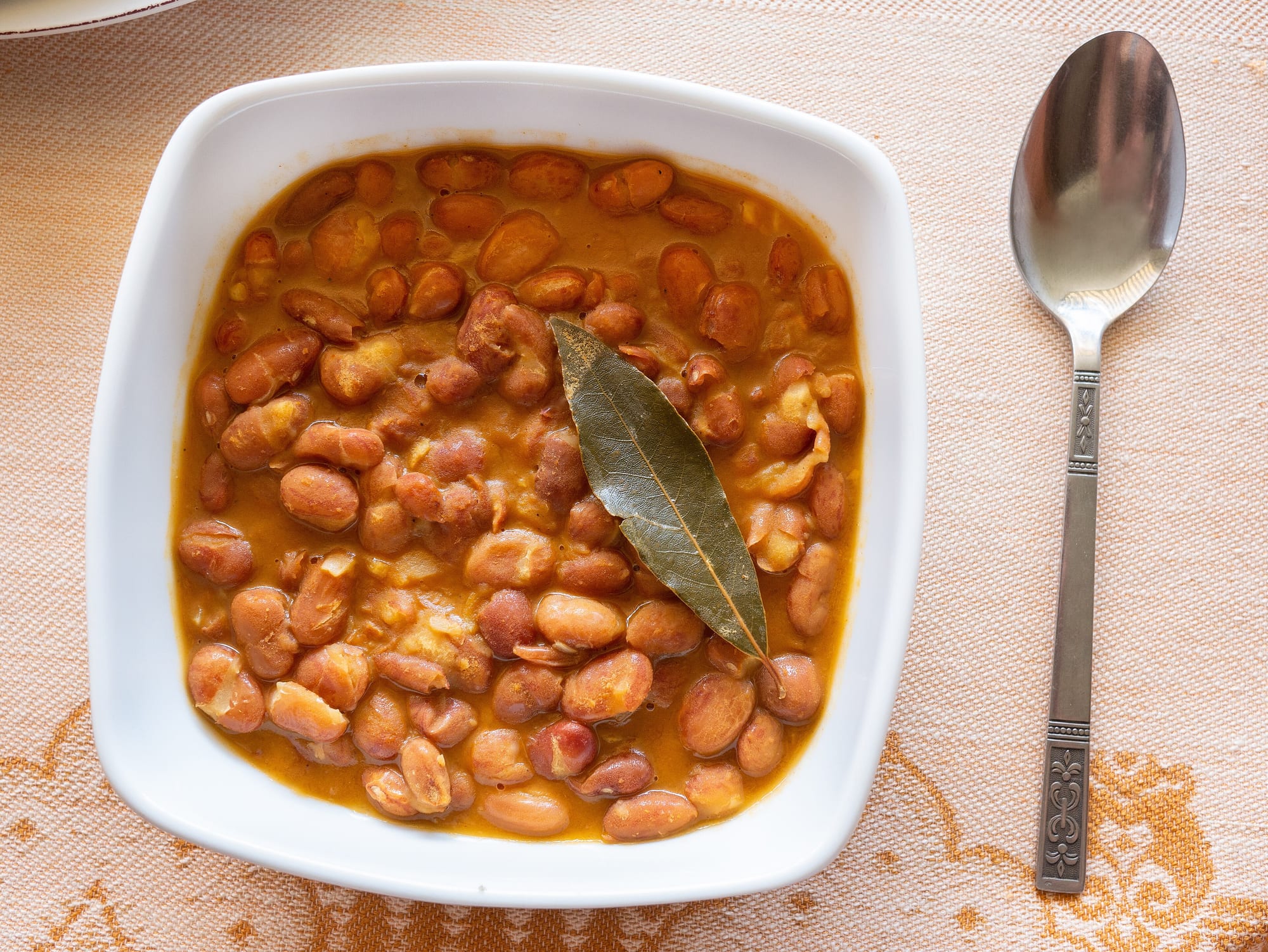 Balkan bean stew of pinto beans in a paprika-red broth with a bay leaf, in a white bowl